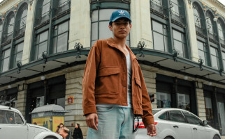Man wearing a brown jacket, blue jeans, and blue cap standing in front of a building