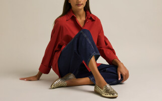 Woman sitting wearing a red blouse, blue jeans, and gold flats