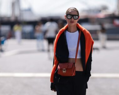 woman stands in street with orange jumper over her shoulders and a crossbody bag